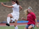 A woman in white in the foreground in midair about to kick a soccer ball while a defensive player readies herself. 